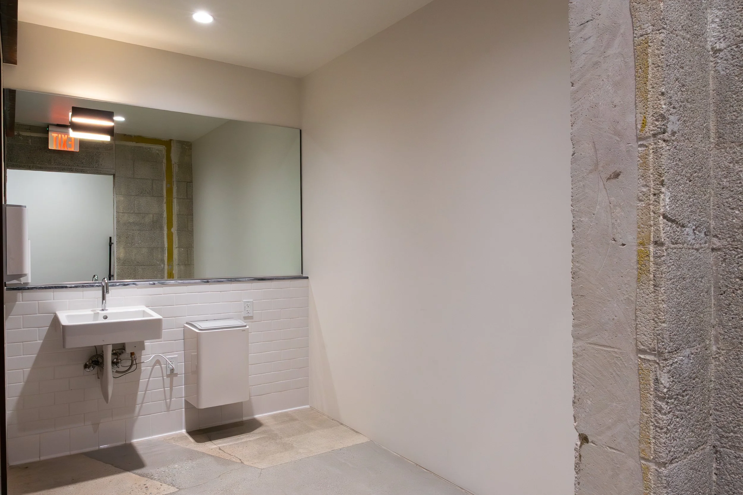 Indoor space with a large mirror, white sink, paper towel dispenser, and exposed brick and concrete wall.
