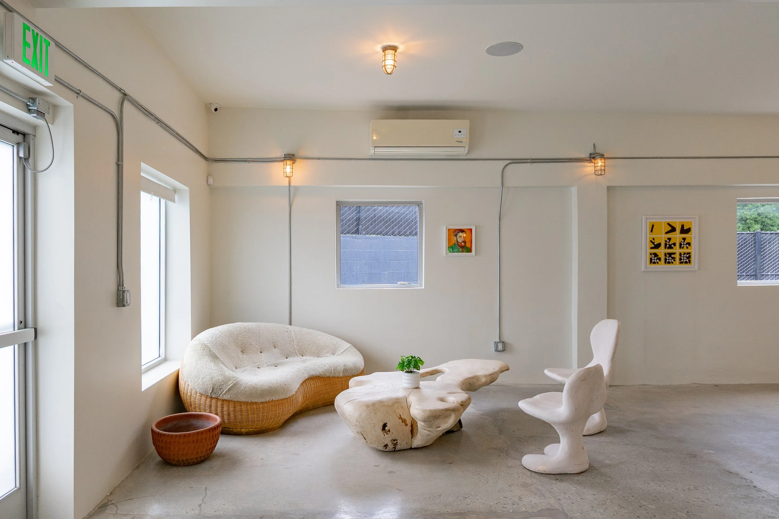 Minimalist interior room with white walls, three small windows, and art on the walls. Contains a wicker sofa with a cream cushion, a uniquely shaped white coffee table with a small potted plant, and two white chairs with abstract forms. There's a red
