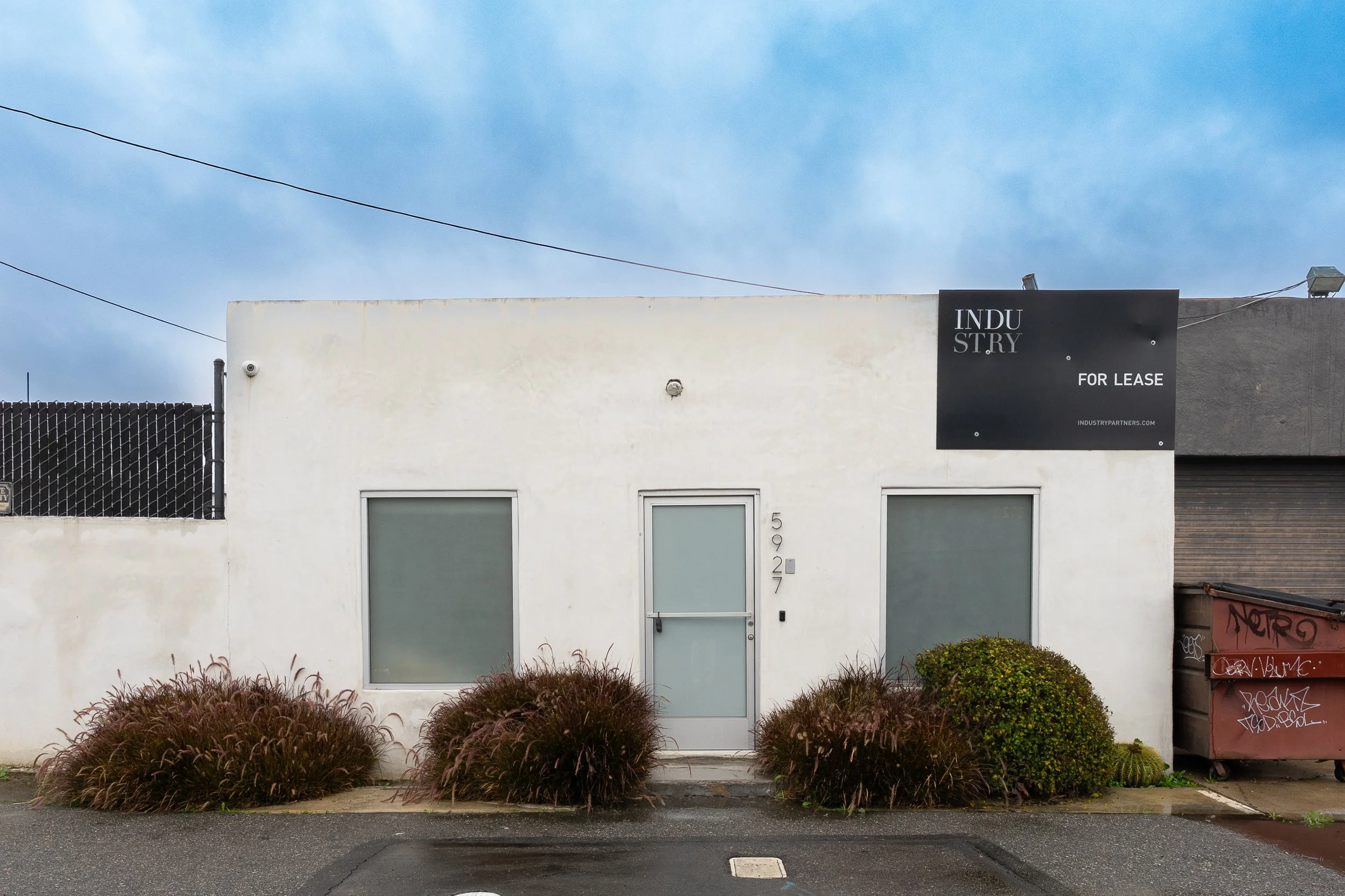 A small white commercial building with a central glass door and two large windows on either side, surrounded by bushes, with a black sign indicating it is available for lease, and a cloudy sky above.