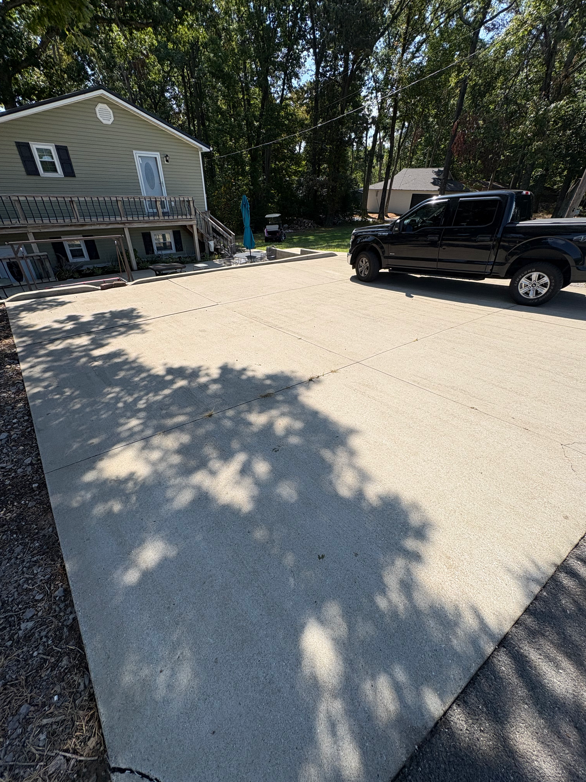 A concrete driveway with a black pickup truck parked on the right. In the background, a house with a balcony, trees, and outdoor furniture. Shadows of trees cast on the driveway.