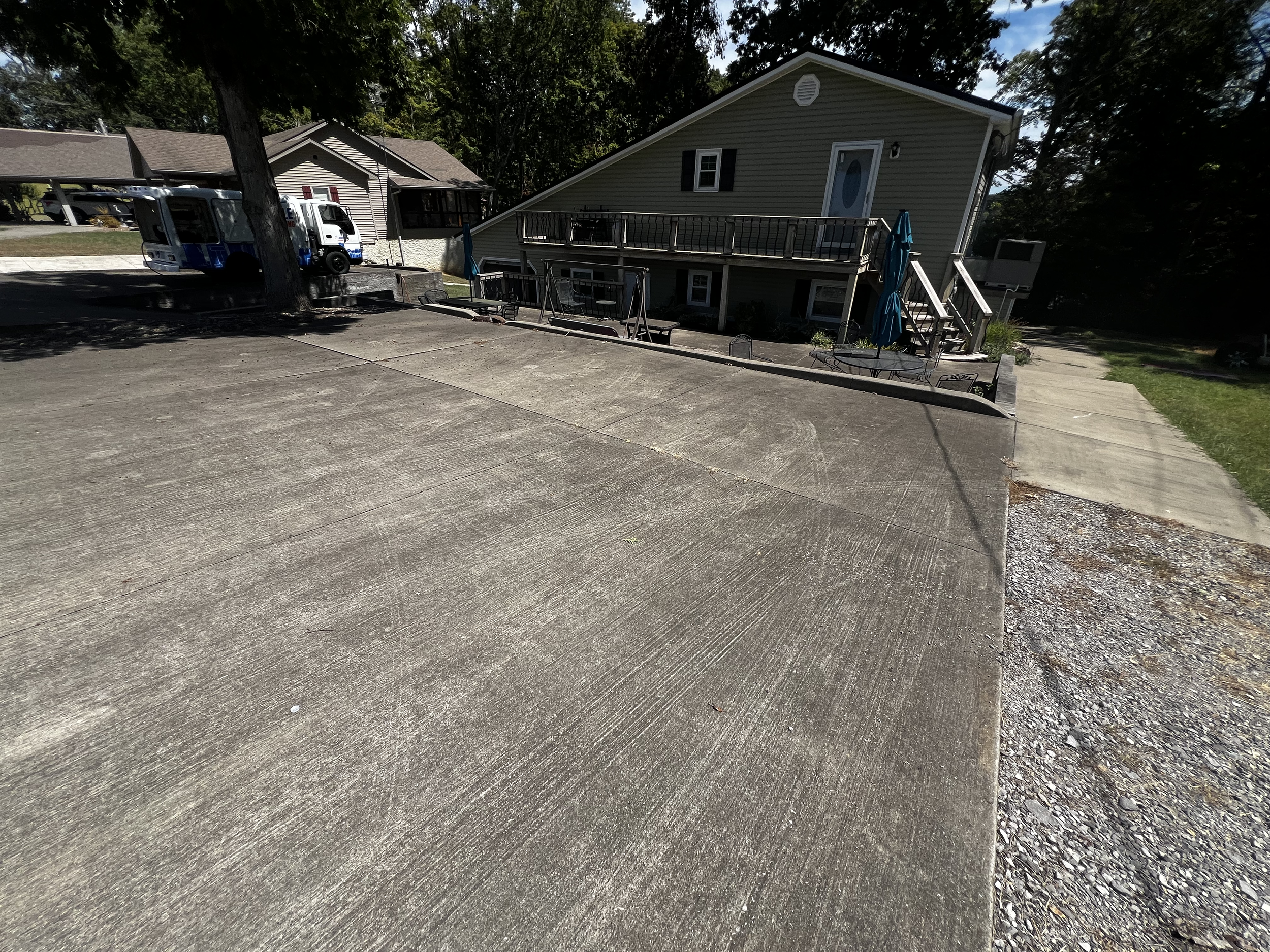 Concrete driveway with visible lines and a gravel area to the right. A house with a wrap-around porch and stairs, surrounded by trees and neighboring houses in the background.