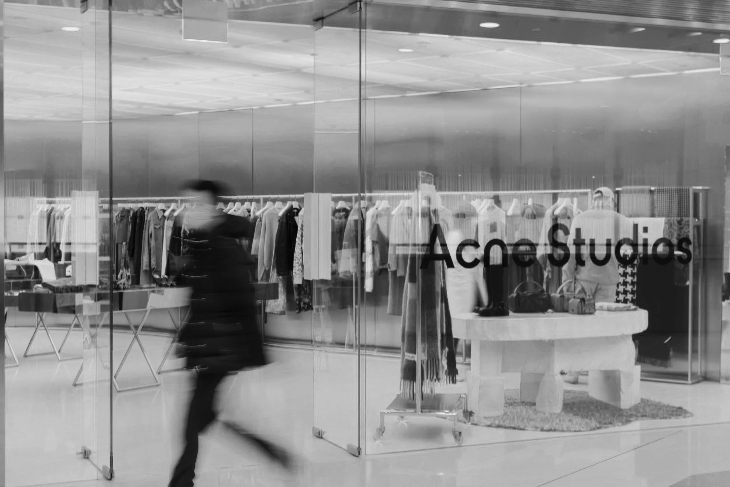 View of a clothing store called Acne Studios with mannequins and racks of clothes inside, seen through glass with the store's name on it, and blurred person walking past.