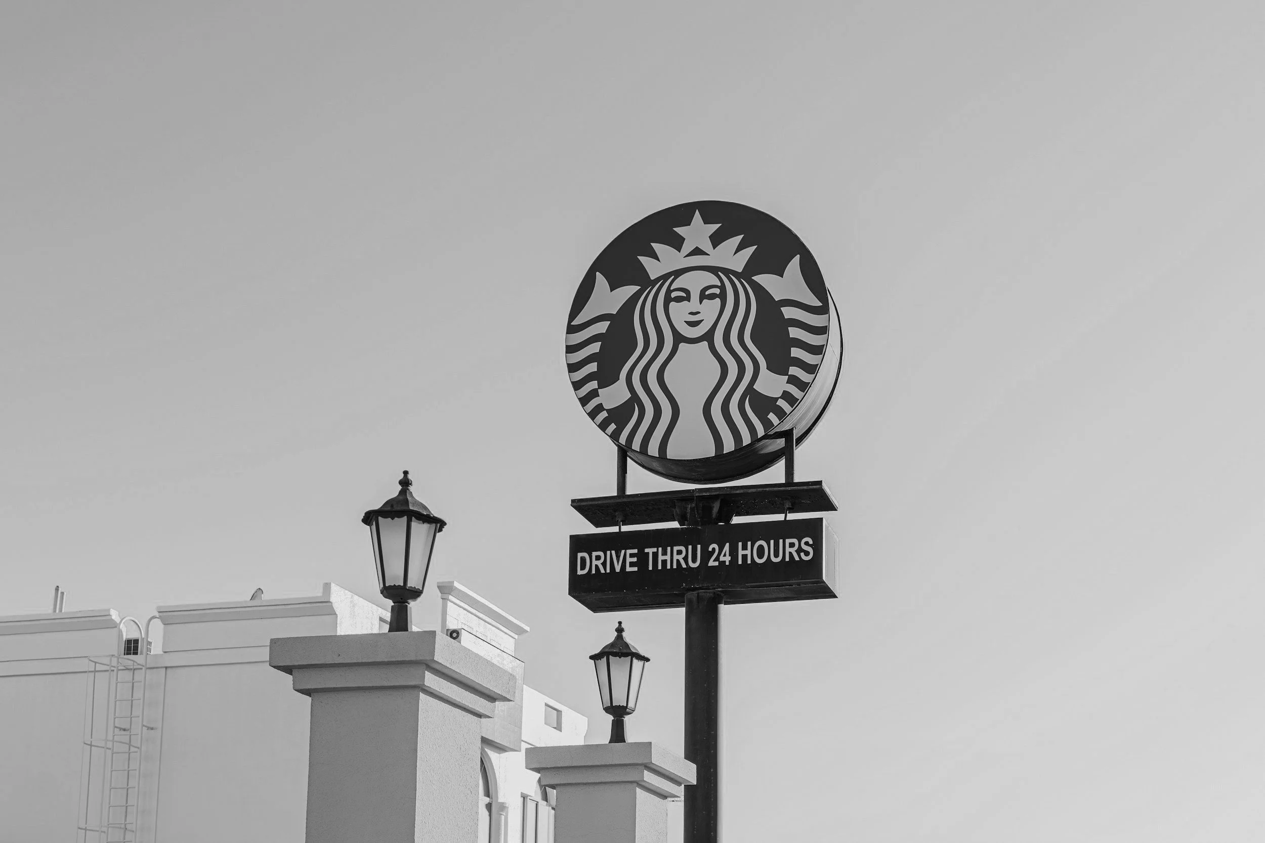 Starbucks coffee shop sign with logo and sign reading "Drive Thru 24 Hours" in black and white.