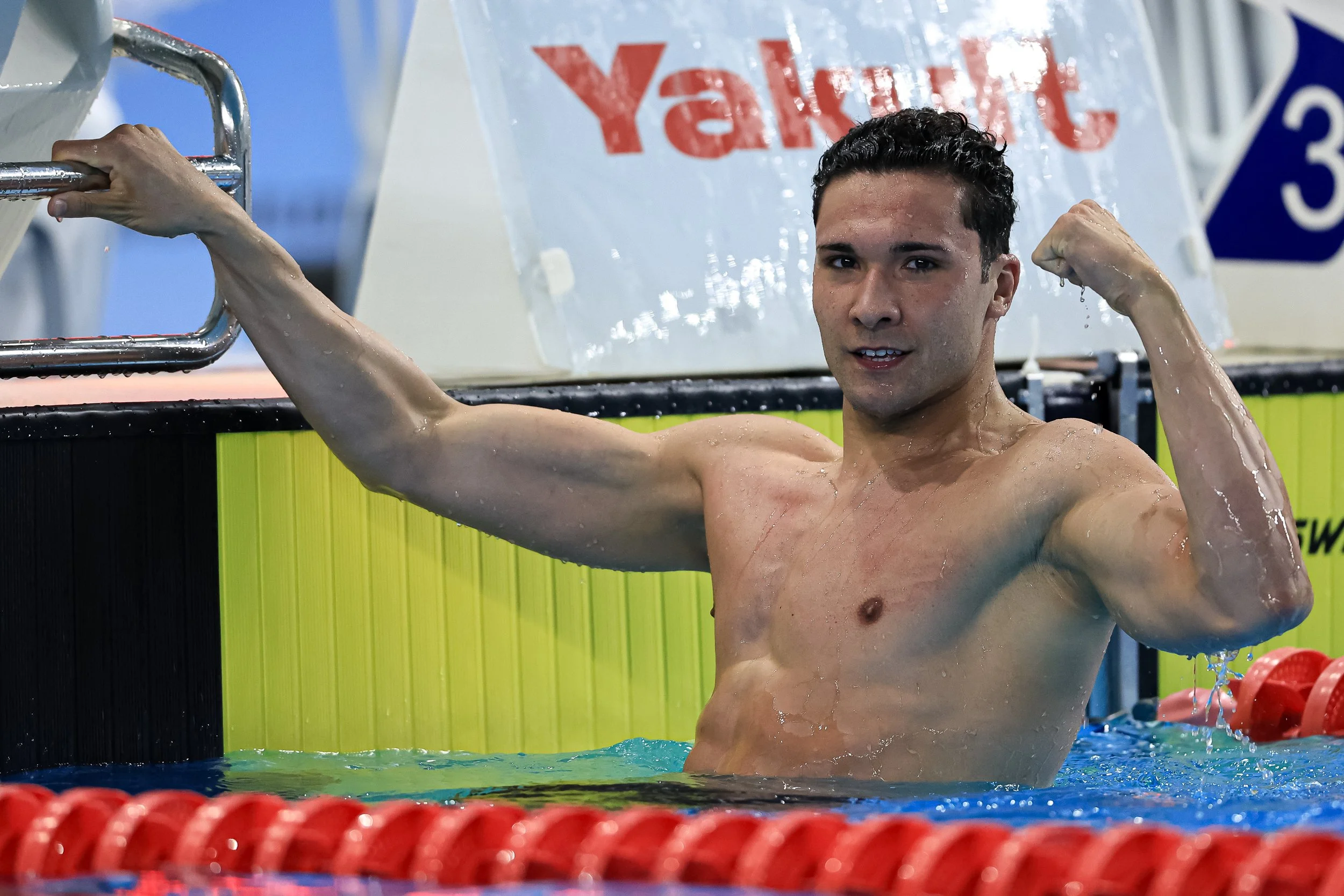 A male swimmer in a swimming pool, flexing his arm and smiling, with wet hair and water droplets on his face and body.