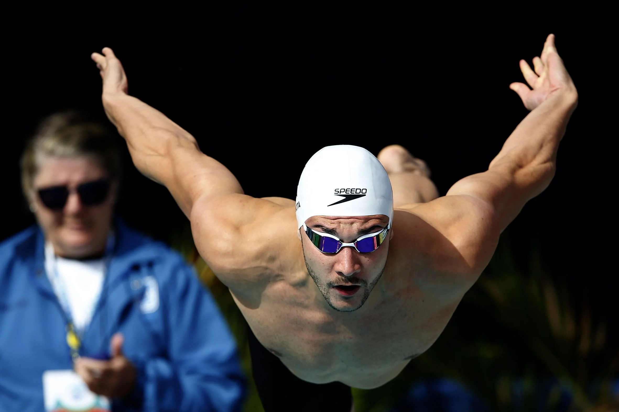 Male swimmer in a diving position wearing goggles, a white swim cap, and no shirt, with a woman in sunglasses and a blue jacket in the background.