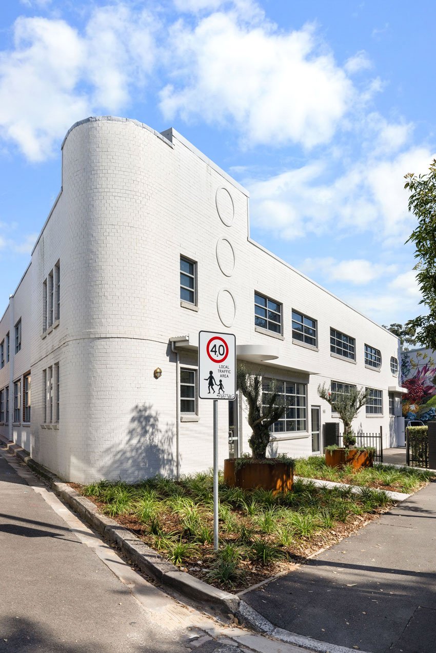 White modern building with multiple windows and a curved corner, street with a 40 mph speed limit sign, landscaped with plants and small trees.