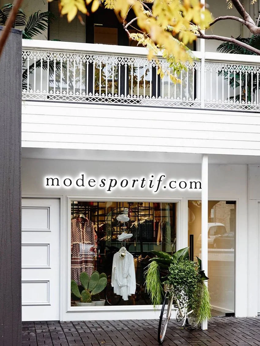Storefront of modesportif.com with window display of clothing and plants, including a cactus and ferns, outside a white building. A bicycle is parked in front of the window.