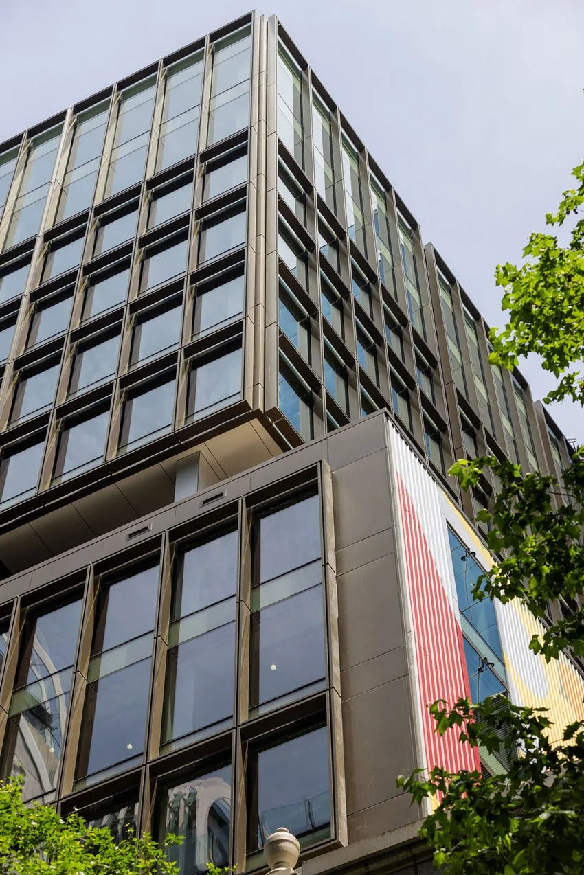 Low-angle view of a modern multi-story building with glass windows, metal framing, and trees nearby.