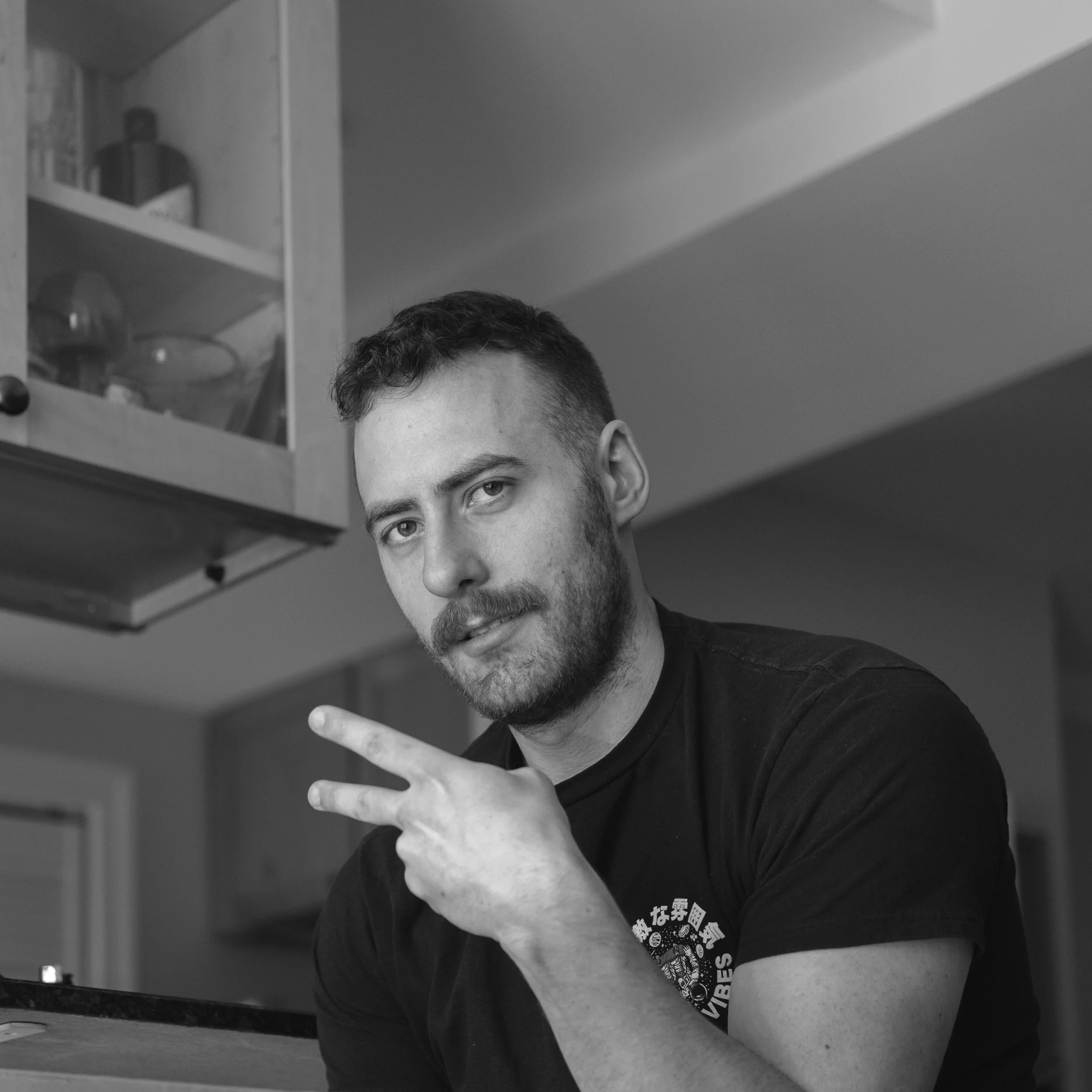 Black and white photo of a young man with short dark hair and a beard, making a peace sign with his right hand while looking at the camera. He's wearing a dark T-shirt and is in a room with a kitchen cabinet in the background.