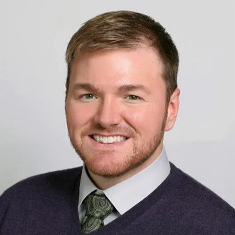 A young man with short brown hair, a beard, and a smile, wearing a white shirt, green tie, and dark sweater, posing against a plain background.