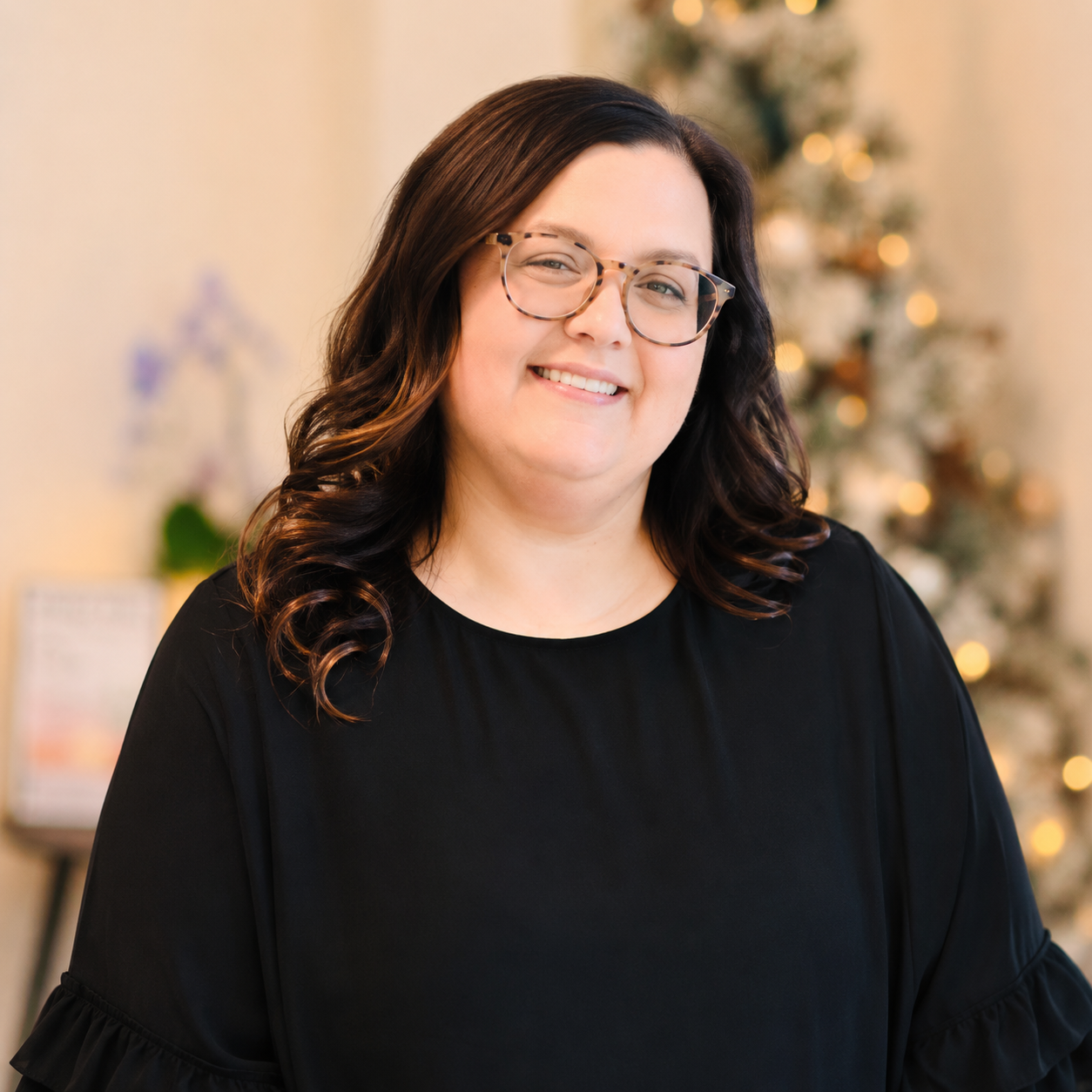 A woman with brown wavy hair and glasses smiling in front of a Christmas tree with lights.