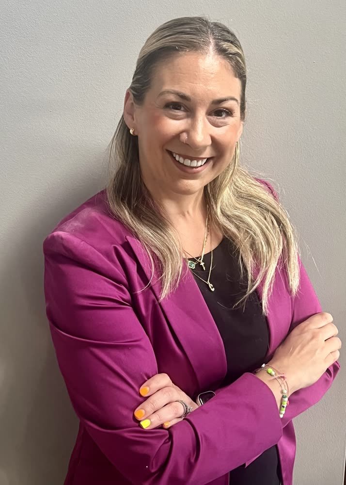 A woman with long, wavy, blonde hair, smiling, wearing a black top, a magenta blazer, and colorful jewelry, standing against a plain gray wall.