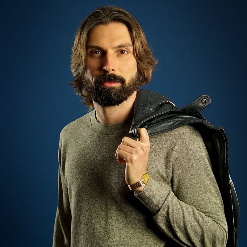 Man with long hair and beard looking directly at camera, wearing a gray shirt, gold watch, and black leather jacket over his shoulder, against a blue background.