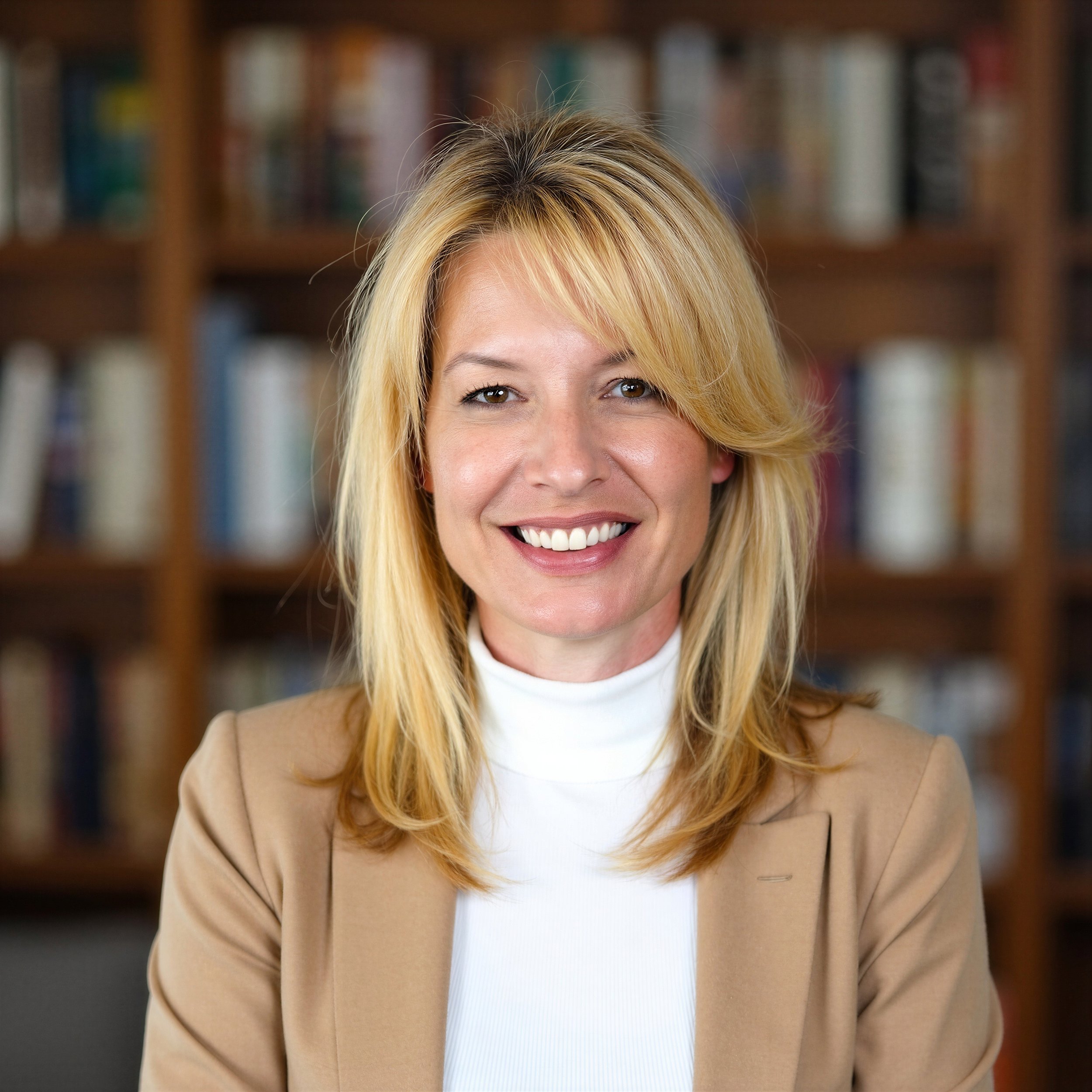 A woman with blonde hair and a beige blazer smiling, with a bookcase in the background.