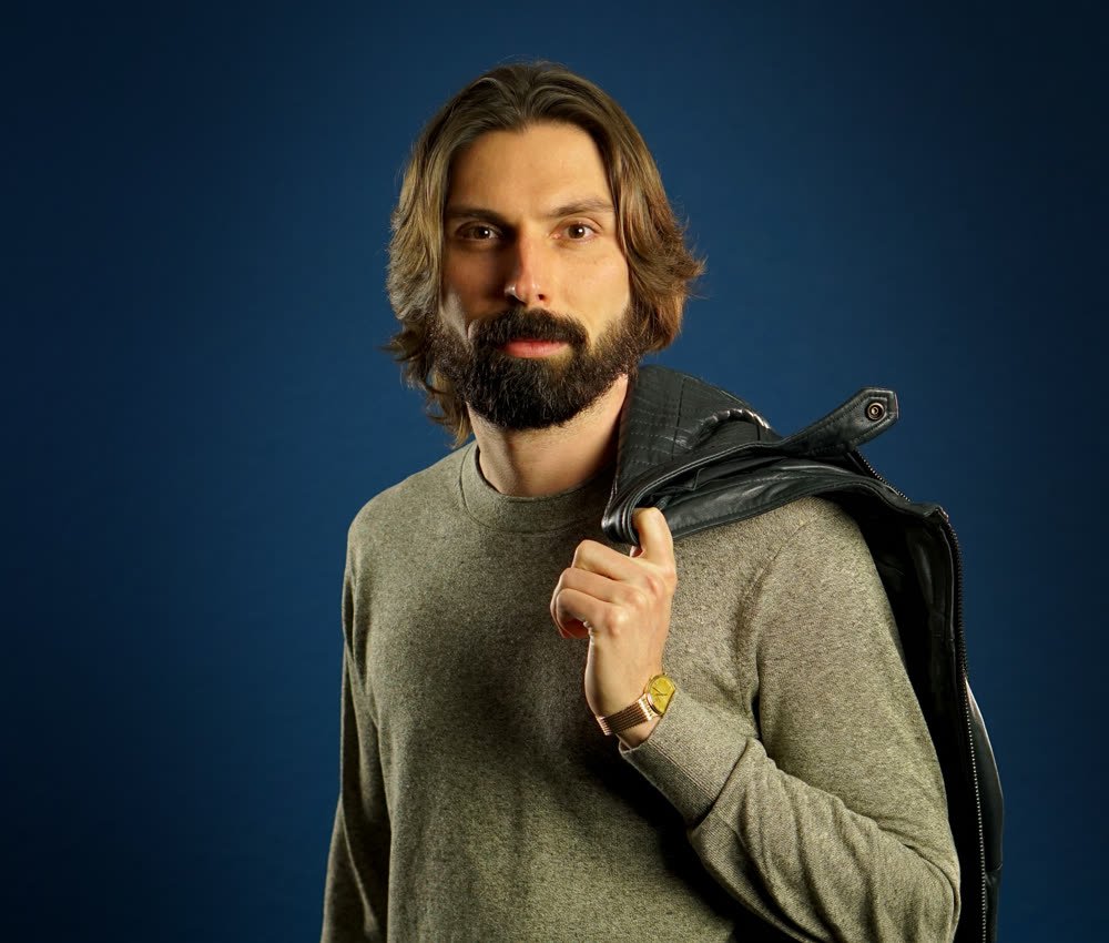 Man with long hair and beard looking directly at camera, wearing a gray shirt, gold watch, and black leather jacket over his shoulder, against a blue background.
