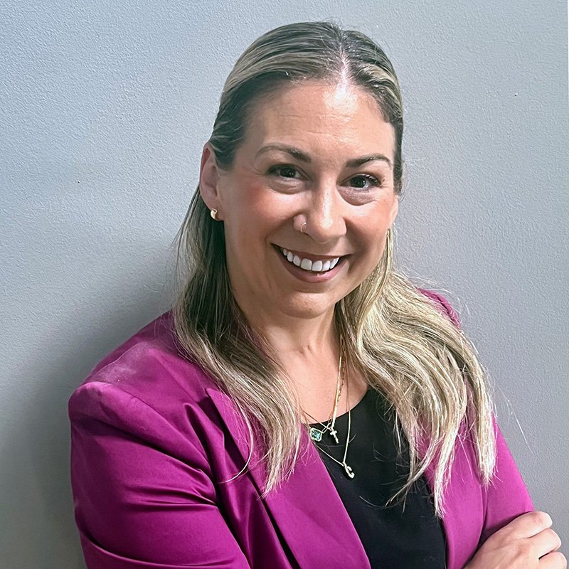 A woman with long, wavy, blonde hair, smiling, wearing a black top, a magenta blazer, and colorful jewelry, standing against a plain gray wall.