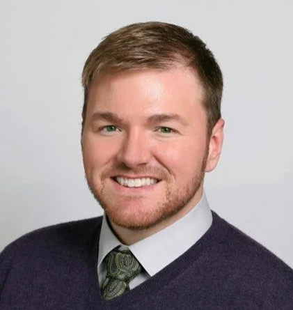 A young man with short brown hair, a beard, and a smile, wearing a white shirt, green tie, and dark sweater, posing against a plain background.