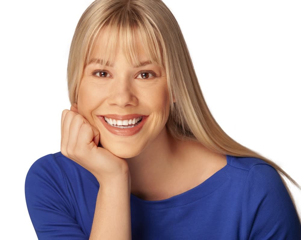 A woman smiling, resting her chin on her hand, wearing a blue top with straight blonde hair and bangs, on a white background.