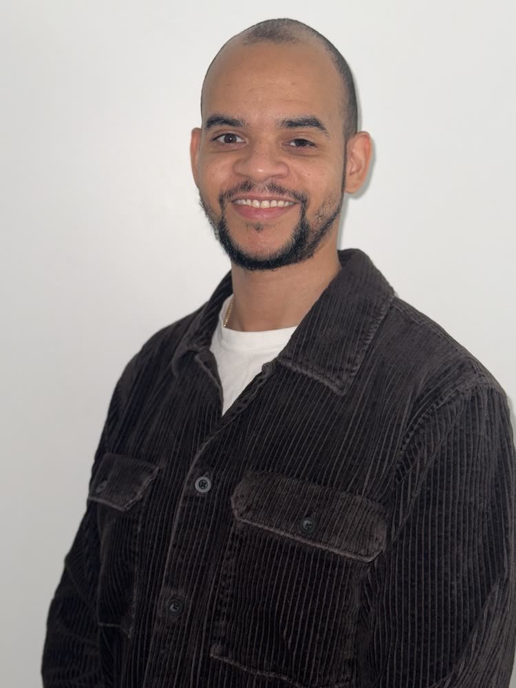 A young man with a beard and mustache, smiling, wearing a dark brown corduroy jacket over a white shirt, standing against a plain white background.