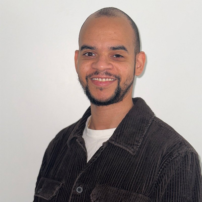A young man with a beard and mustache, smiling, wearing a dark brown corduroy jacket over a white shirt, standing against a plain white background.