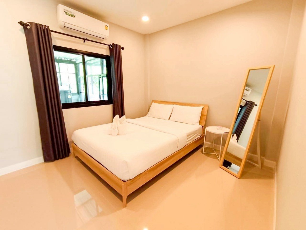Hotel room with a wooden bed frame, white bedding, a bedside table, a full-length mirror, a window with dark curtains, and an air conditioning unit