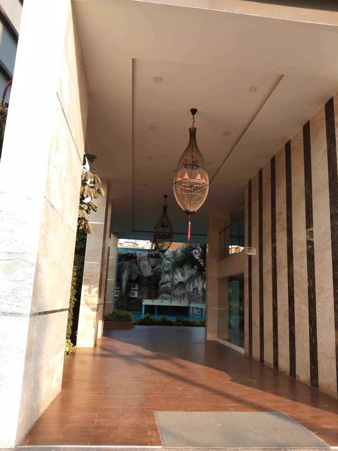 Elegant corridor with hanging chandeliers, stone and wood wall accents, leading to an outdoor area with rocky sculpture and palm trees.