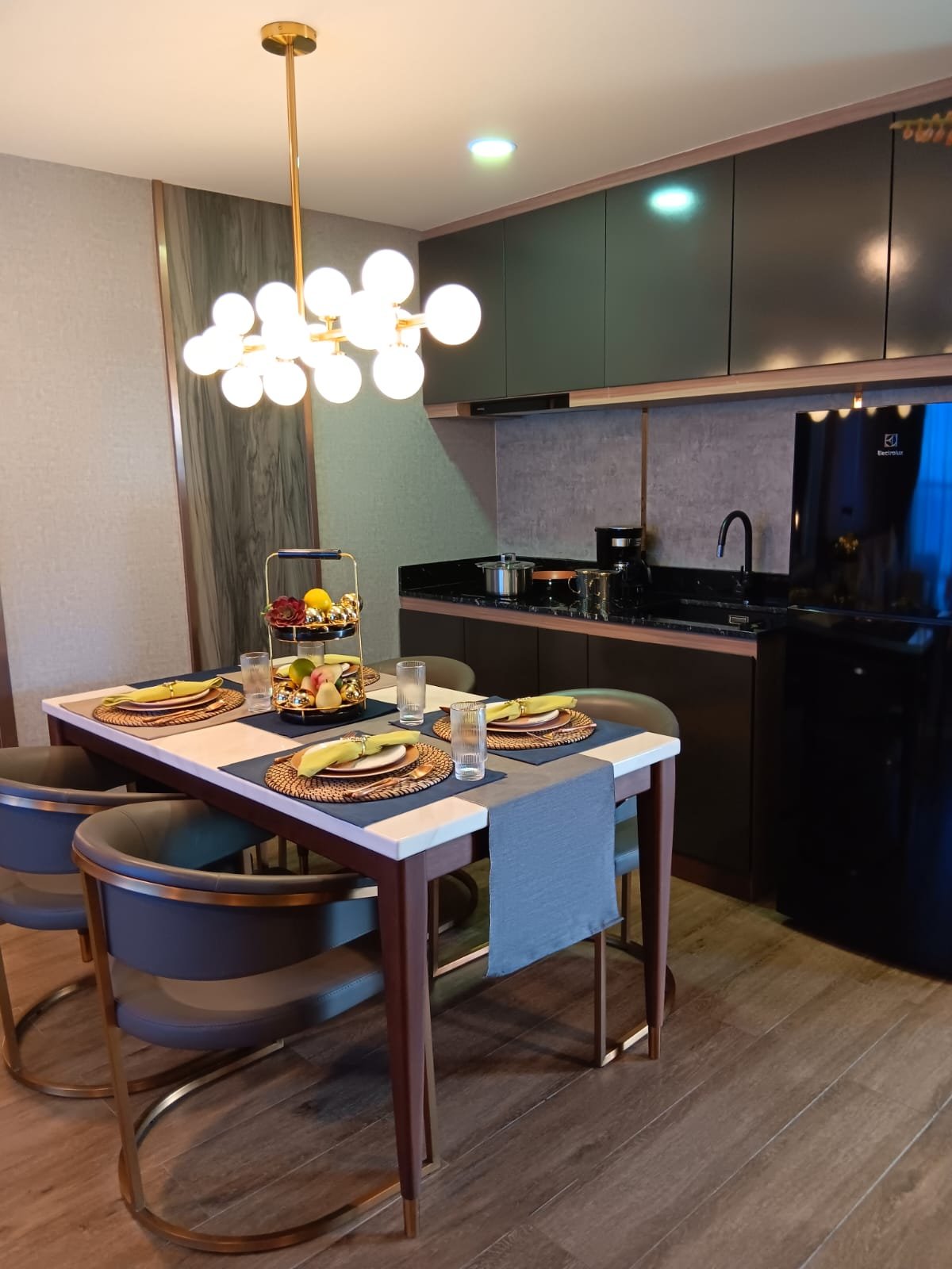 Modern dining area with a white table set for four, featuring yellow napkins, woven placemats, and glassware, under a chandelier with round bulbs, next to a black kitchen with green cabinets and a black refrigerator.