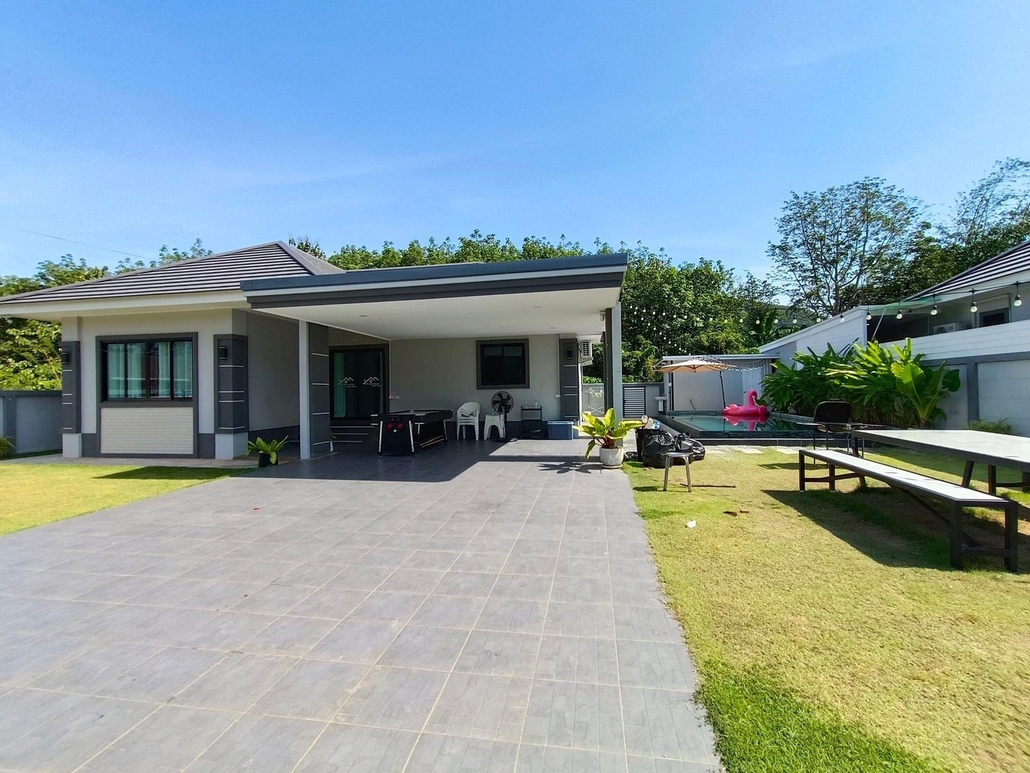 Modern house with a grey tile patio, outdoor seating, small pool with inflatable float, green plants, and string lights on a sunny day.