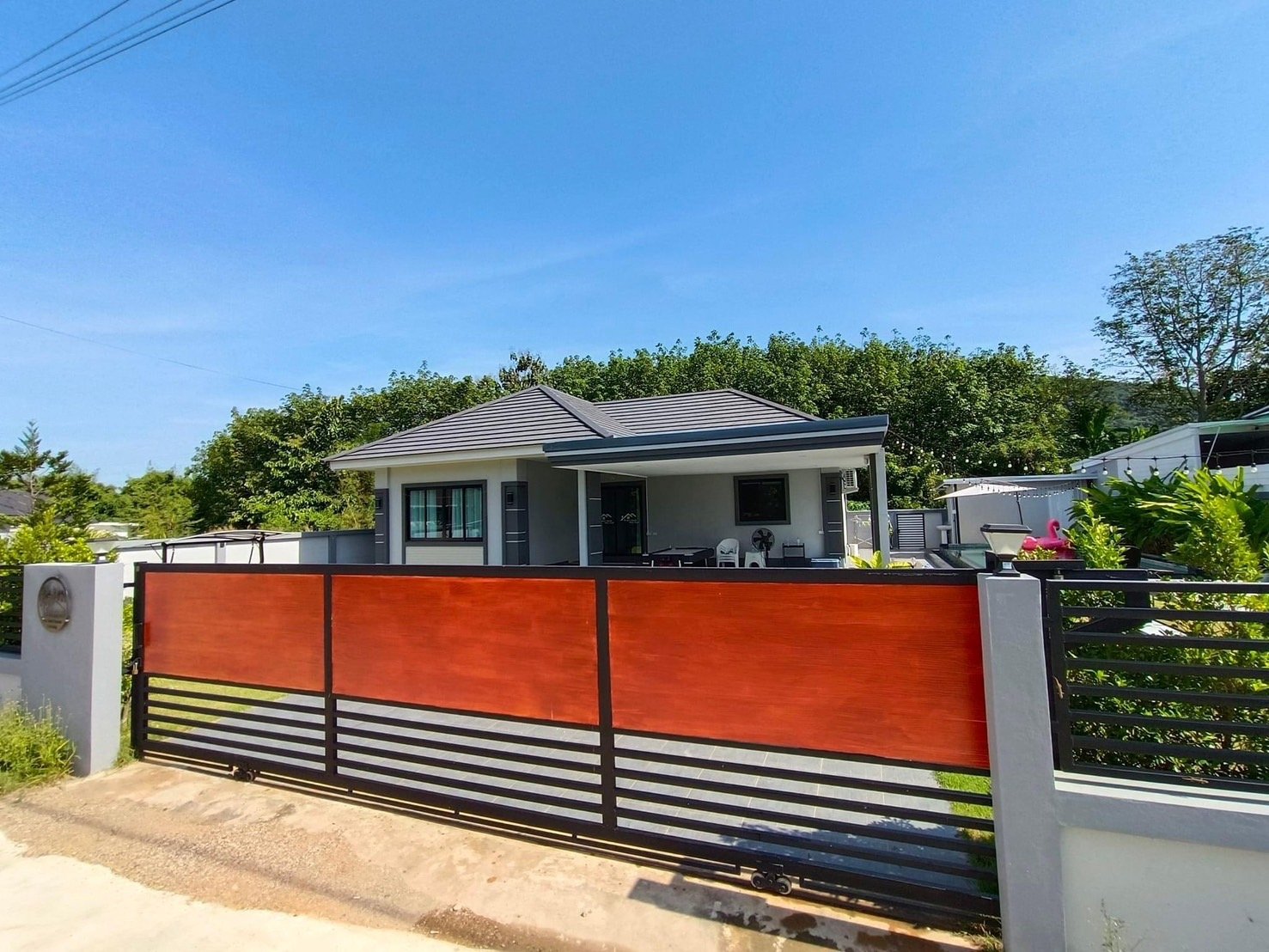 Modern single-story house with gray walls, black window frames, and a gray tiled roof, surrounded by a black and orange gate, greenery, and a clear blue sky.