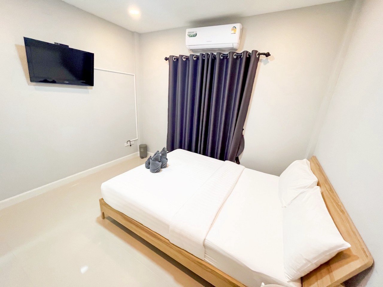 A minimalist hotel room with a bed, a wall-mounted TV, an air conditioning unit, dark curtains, and a pair of gray slippers on the bed.