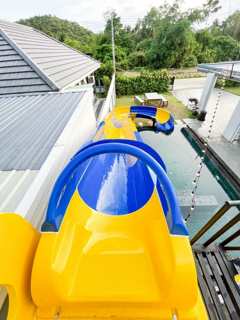 Colorful yellow and blue water slide leading to a swimming pool in a backyard outdoor setting with trees and greenery in the background.