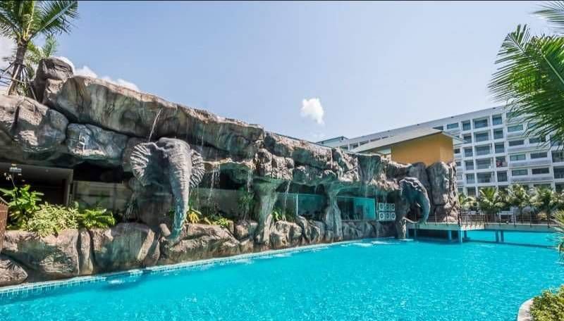 A hotel pool area with a large artificial rock wall featuring elephant sculptures and water spouts, surrounded by palm trees and a multi-story hotel building in the background.