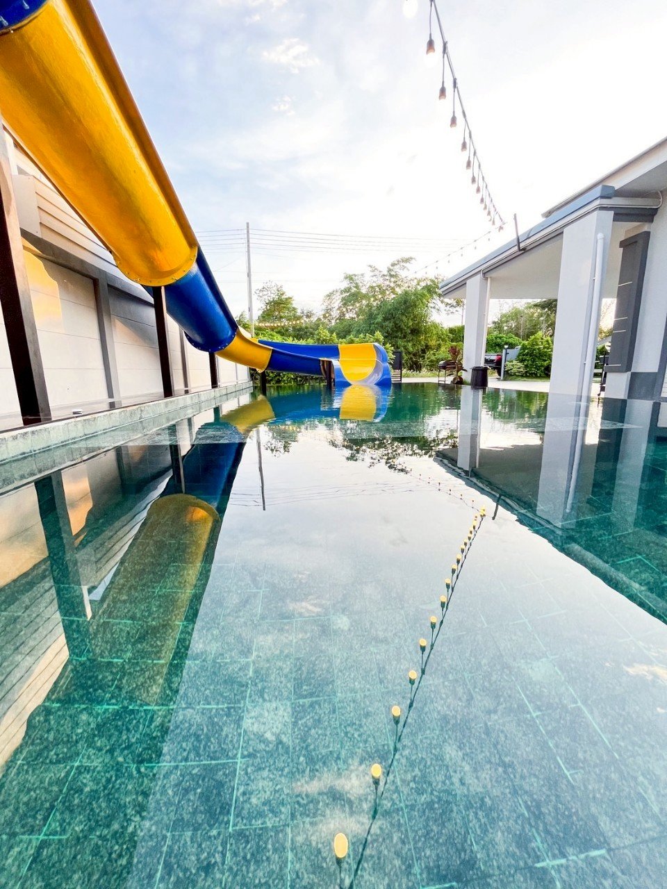A swimming pool with a yellow and blue water slide, outdoor string lights, and surrounding trees and houses.