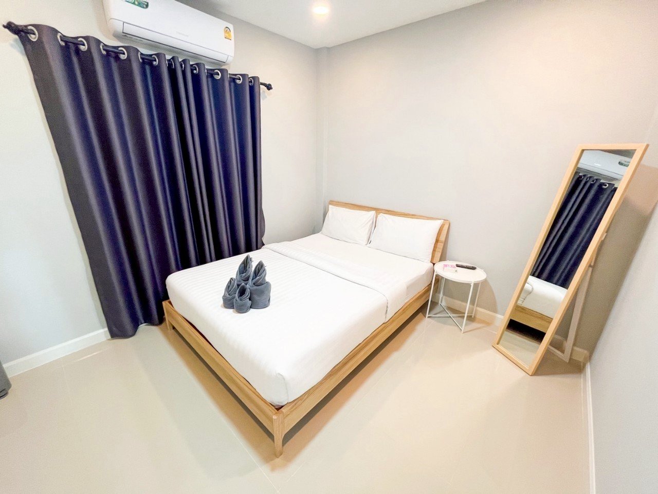Minimalist bedroom with a wooden bed frame, white bedding, three rolled grey towels on the bed, a small white round side table with a remote control, a large standing mirror with a wooden frame, a dark curtain covering a window, and a wall-mounted ai