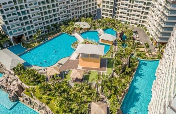 Aerial view of a resort pool area with multiple swimming pools, surrounded by tall residential buildings, palm trees, and lounge areas.