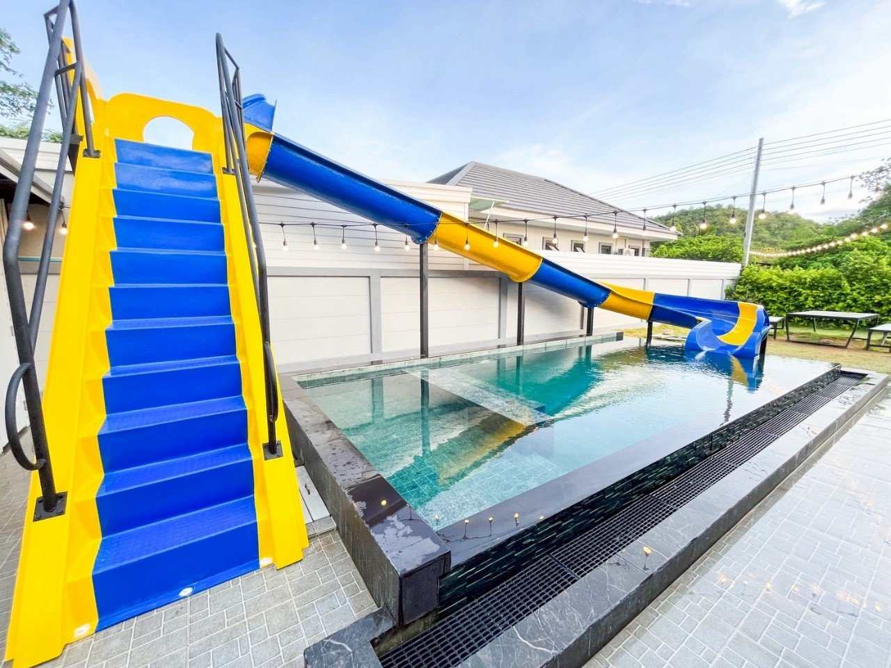 An outdoor swimming pool with a blue and yellow waterslide and a pool deck, surrounded by a white fence and string lights.