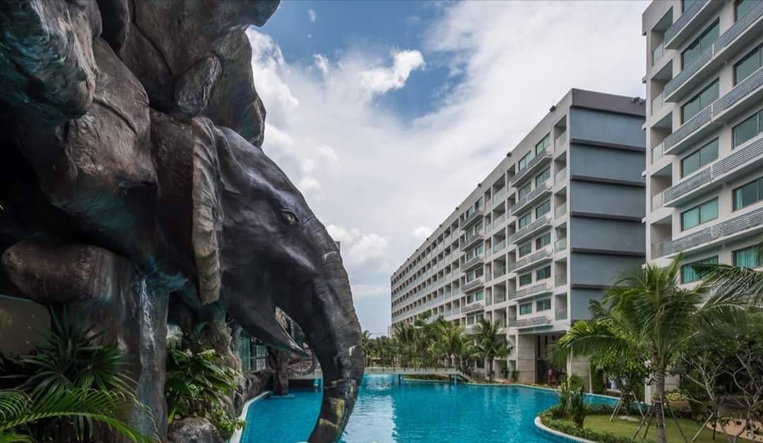 A hotel pool area with a large elephant sculpture on the side, surrounded by tropical plants and modern multi-story buildings, under a partly cloudy sky.
