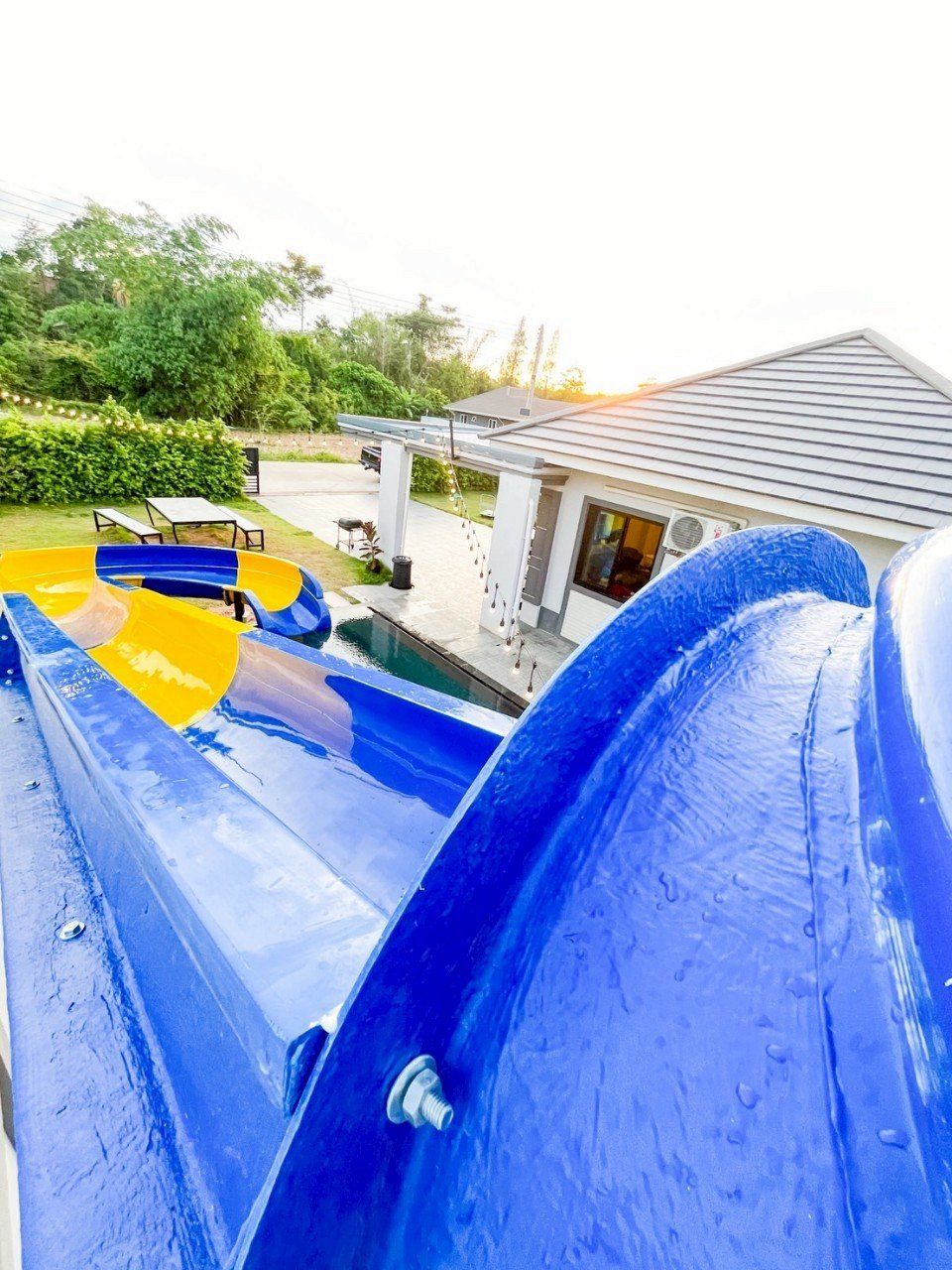A colorful water slide in a backyard with a pool, surrounded by outdoor furniture and greenery, during sunset.
