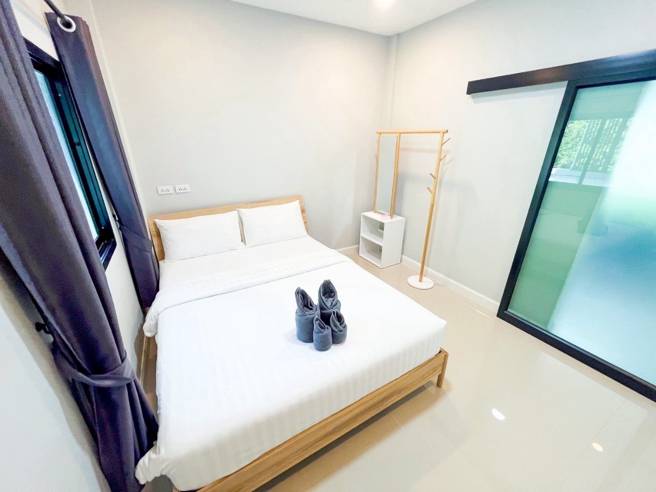Minimalist bedroom with a bed, folded towels, gray curtains, a small white shelf, wooden clothing rack, and large window with frosted glass.