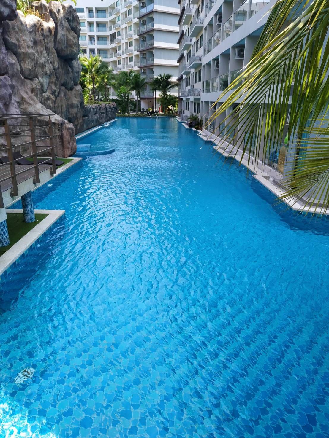 Swimming pool between tall apartment buildings, with a rocky wall and palm trees in the background.