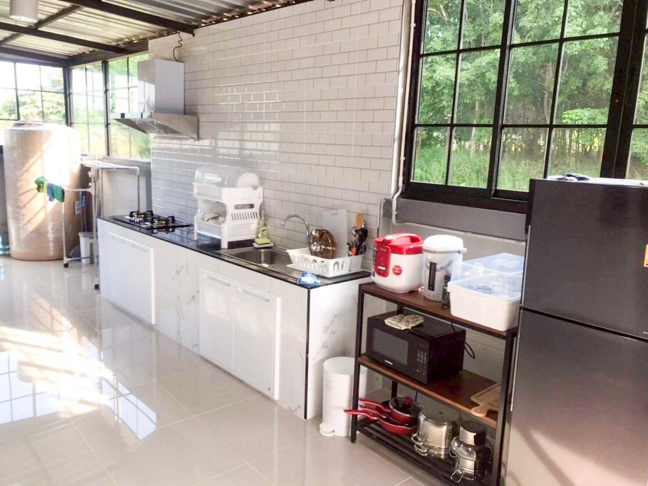 Kitchen with white tiled walls, large window showing greenery, microwave, refrigerator, rice cooker, and various kitchen appliances and utensils.