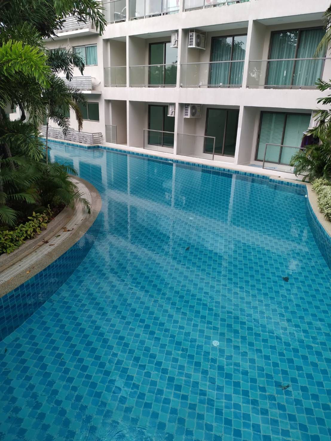 Swimming pool surrounded by apartment building with glass doors and balconies, greenery on the side.