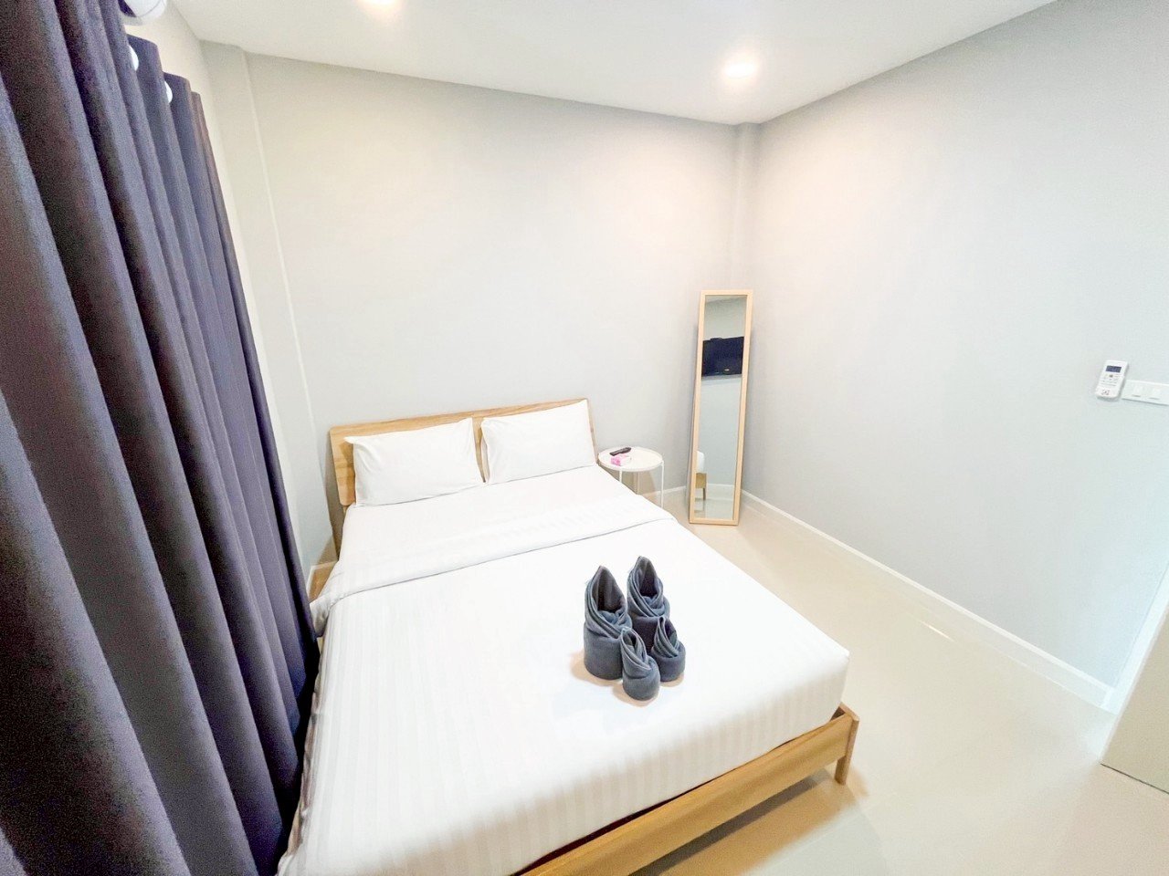Minimalist hotel bedroom with a wooden bed frame, white bedding, pillows, a pair of gray slippers on the bed, a small round bedside table, a full-length mirror, dark curtains, and a wall-mounted thermostat.