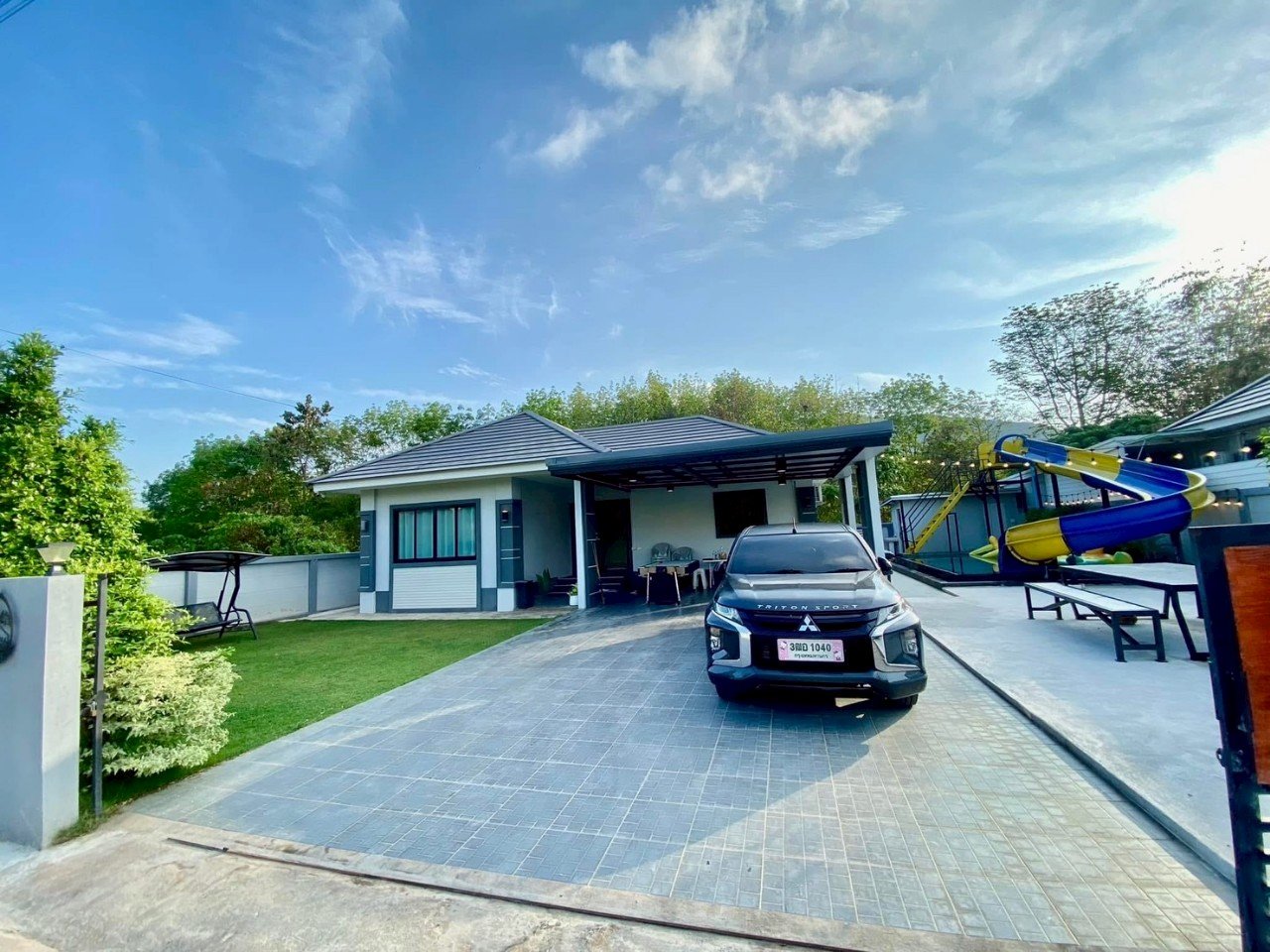 Modern house with gray exterior, parked black Mitsubishi Triton Sport, colorful outdoor water slide, patio, green lawn, and trees under a blue sky.