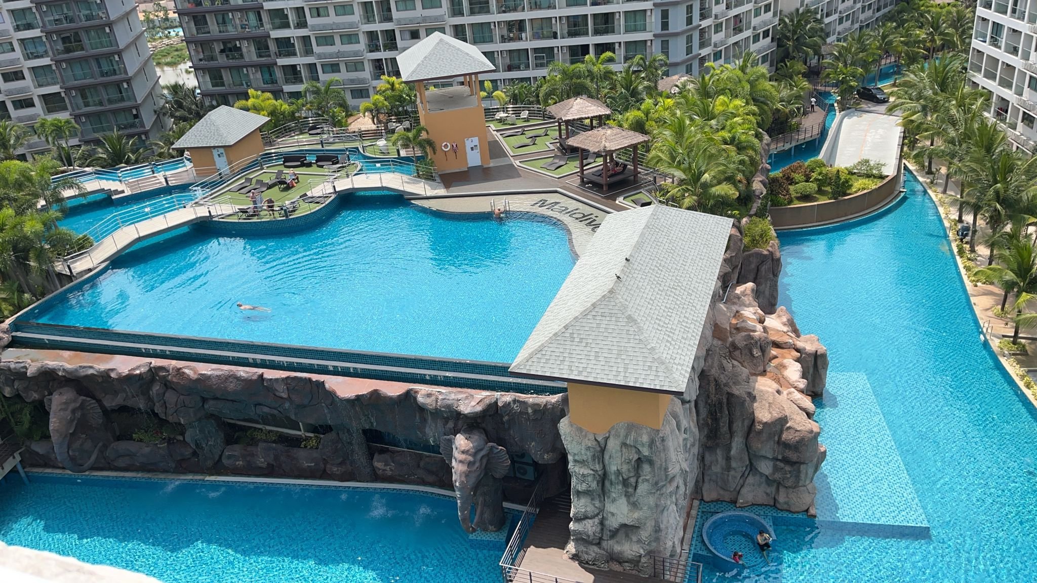 A large outdoor swimming pool area at a resort with surrounding high-rise buildings. The pool has a cartoonish design with rocky elements, water slides, and shaded lounging areas. Lush green palm trees and tropical plants decorate the landscape.