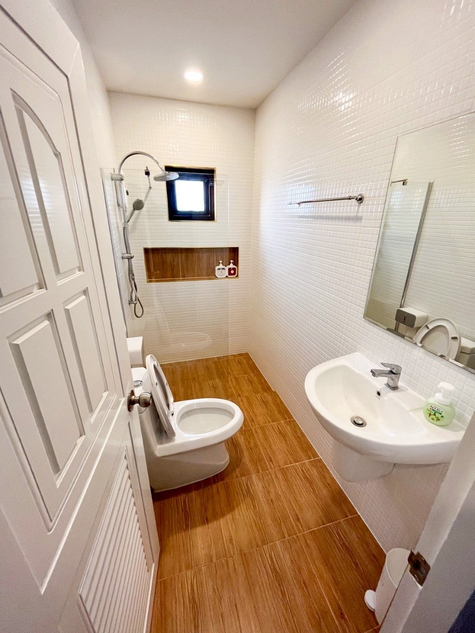 Small bathroom with white tiled walls, wooden floor tiles, a small window, a showerhead, a toilet, a wall-mounted sink, a mirror, and a small built-in shelf with bottles.