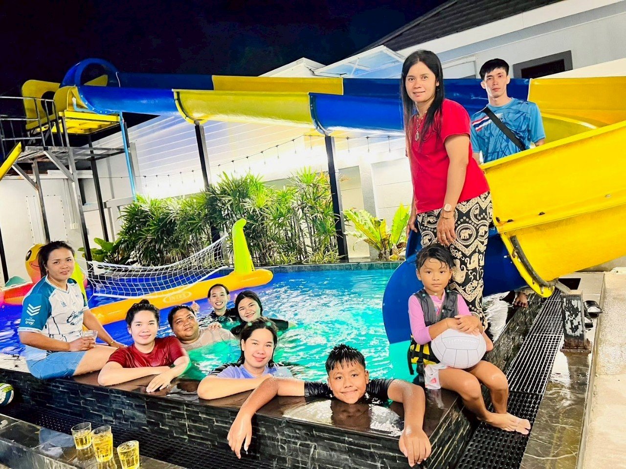 A group of people enjoying a night swim at a water park, with a water slide and pool, surrounded by plants and a white building.