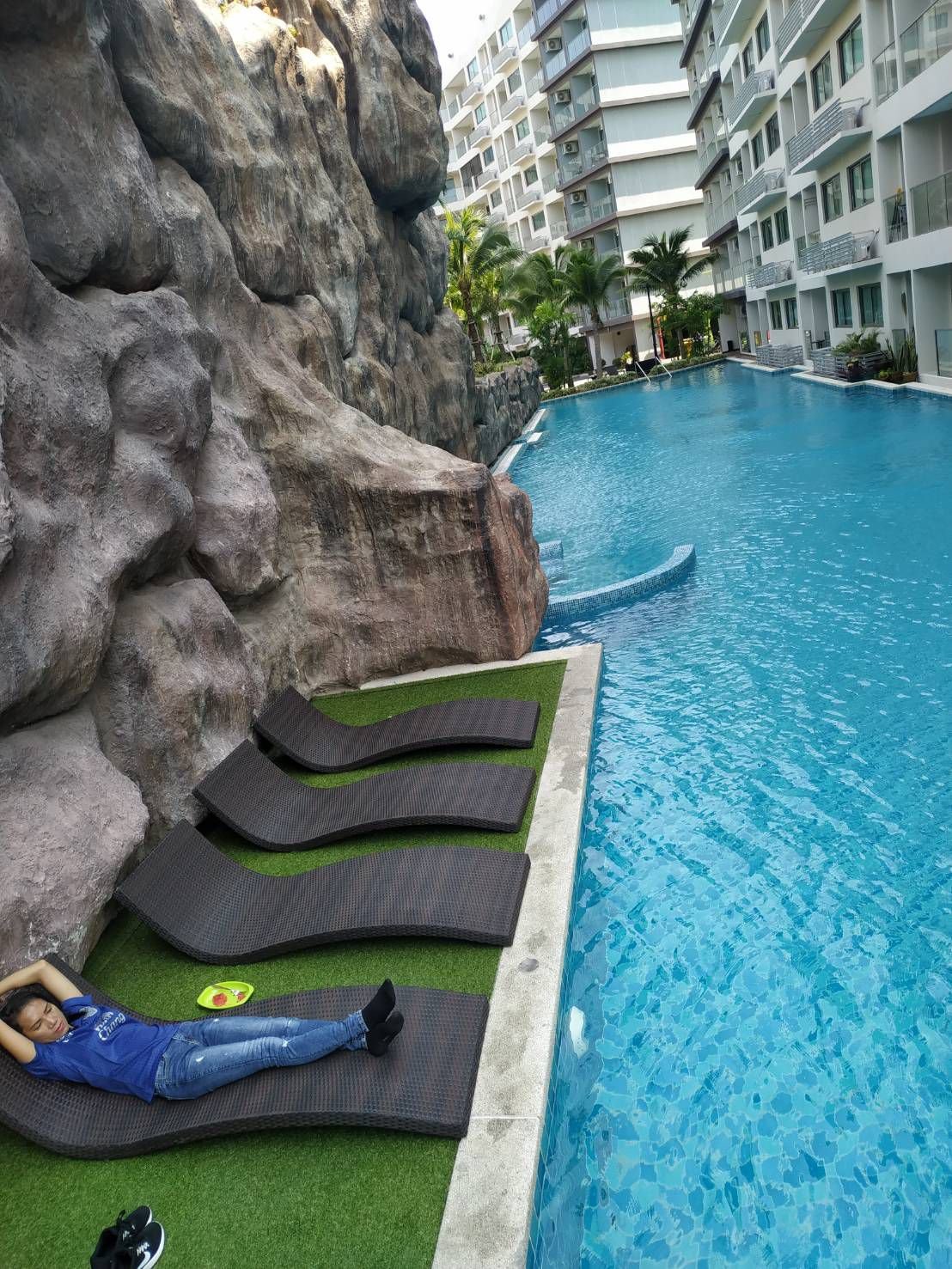 A woman lying on a black lounge chair beside a swimming pool with rock formations on the left and modern apartment buildings in the background.