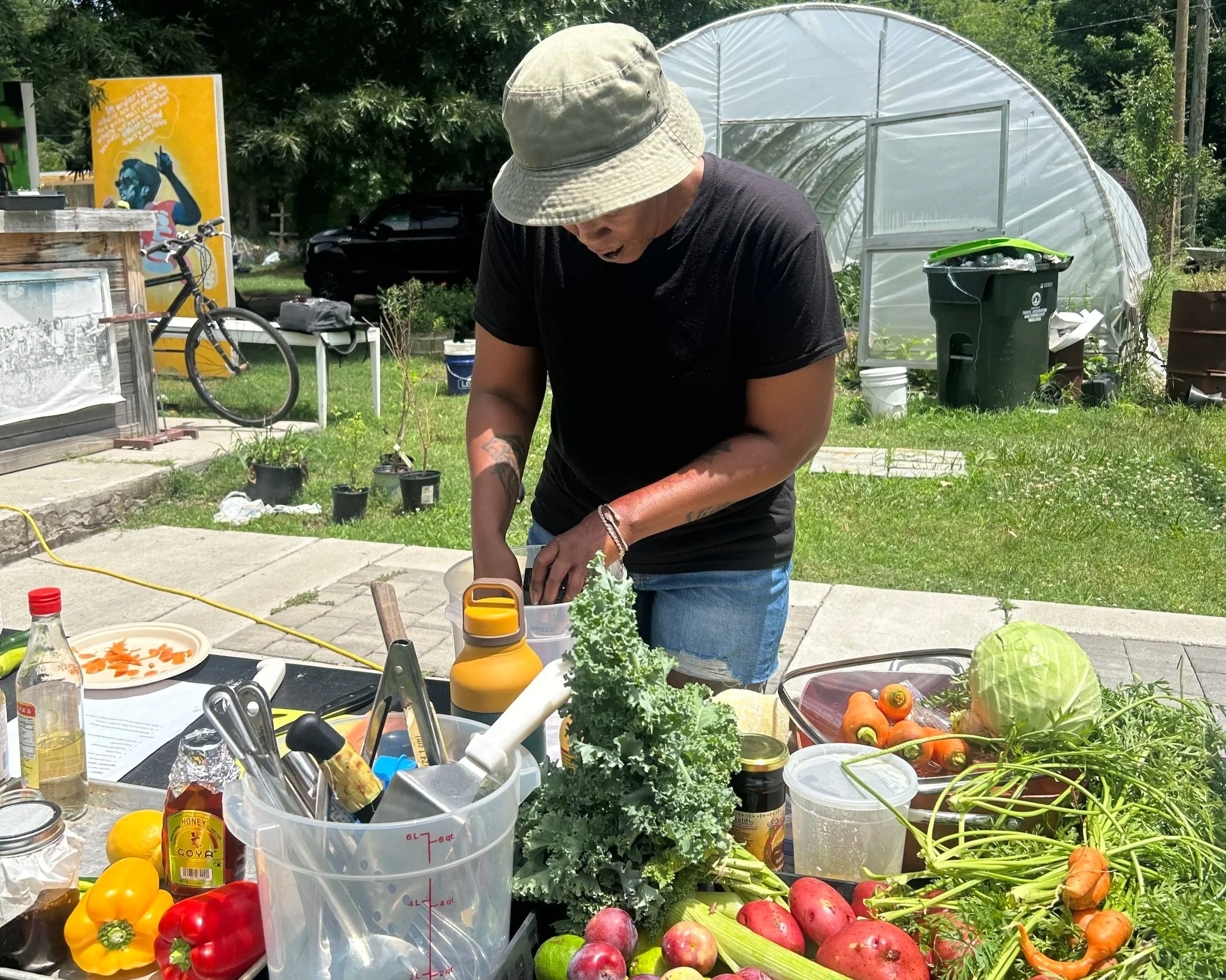 A man wearing a beige bucket hat and black t-shirt is working outdoors at a garden table filled with various vegetables and gardening supplies. There is a greenhouse, a bike, and trees in the background.