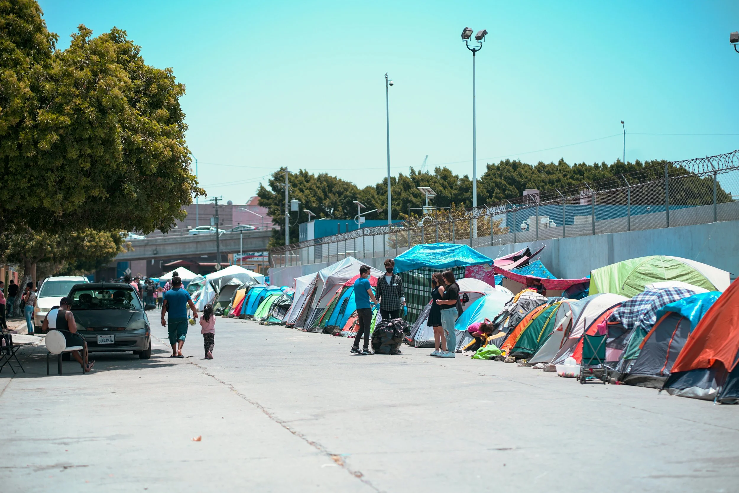 People standing and sitting near tents and cars in an outdoor setting on a sunny day, with a tree on the left and streetlights overhead.